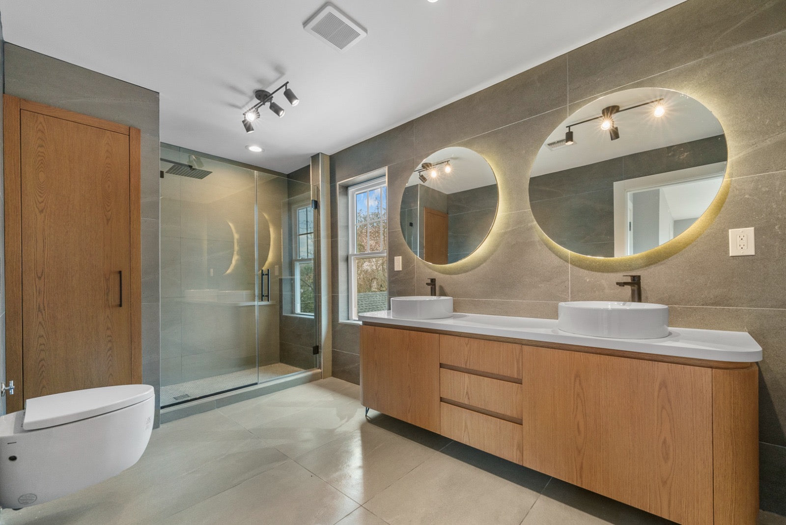 Two illuminated round mirrors above a wood double vanity